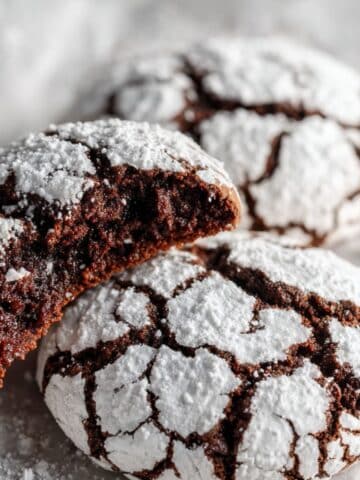 Homemade chocolate crinkle cookies with white powdered sugar cracks, one bitten showing fudgy interior Three chocolate crinkle cookies on parchment paper displaying perfect cracking and brownie-like texture
