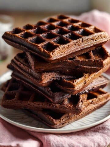 Overhead shot of thick, fudgy brownie waffles stacked on a vintage dessert plate, with crisp waffle edges and rich chocolate texture, styled on a rustic table with a pastel napkin for a cozy homemade look.