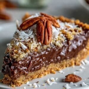 Slice of no-bake German chocolate pie with fudgy chocolate filling and gooey coconut pecan topping. Golden graham crust and homemade texture on white plate.