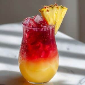 Refreshing Malibu Bay Breeze cocktail in a curvy glass on a white marble counter, showcasing vibrant layers of pineapple yellow and cranberry red, topped with crushed ice and garnished with a fresh pineapple wedge; condensation on the glass enhances the tropical, summery appeal.