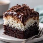 Close-up side view of a homemade Mounds cake slice with a moist chocolate base, thick coconut layer, and glossy chocolate ganache topping, finished with rich chocolate shavings on a ceramic plate.