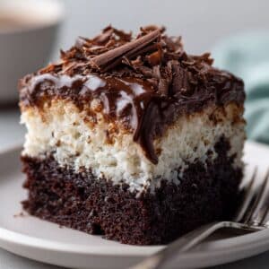 Close-up side view of a homemade Mounds cake slice with a moist chocolate base, thick coconut layer, and glossy chocolate ganache topping, finished with rich chocolate shavings on a ceramic plate.