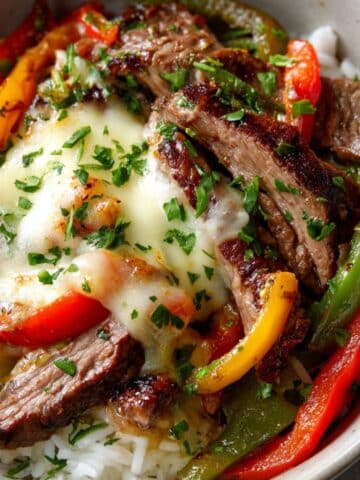 Close-up of a Philly cheesesteak bowls topped with gooey provolone cheese, tender seared steak strips, sautéed bell peppers, and fresh parsley, all served over fluffy white rice in a homemade, comforting presentation.