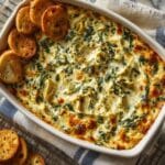 Creamy spinach artichoke dip baked to perfection in a white casserole dish, topped with melty cheese and surrounded by golden crostini crackers.