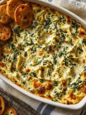 Creamy spinach artichoke dip baked to perfection in a white casserole dish, topped with melty cheese and surrounded by golden crostini crackers.