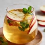 Refreshing apple cider vinegar drink in a clear glass, filled with apple slices, fresh mint, and sparkling golden liquid on a wooden coaster.