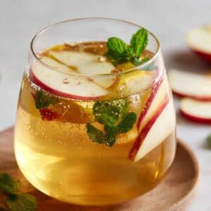 Refreshing apple cider vinegar drink in a clear glass, filled with apple slices, fresh mint, and sparkling golden liquid on a wooden coaster.
