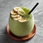 Creamy banana matcha smoothie in a round glass, topped with sliced bananas and matcha powder, served with a black straw on a wooden coaster over gray marble countertop.