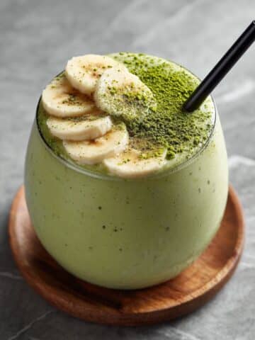 Creamy banana matcha smoothie in a round glass, topped with sliced bananas and matcha powder, served with a black straw on a wooden coaster over gray marble countertop.