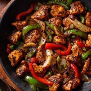 Juicy black pepper chicken stir-fry cooked in a skillet with glossy sauce, tender chicken bites, red and green bell peppers, and caramelized onions tossed in bold cracked black pepper seasoning.