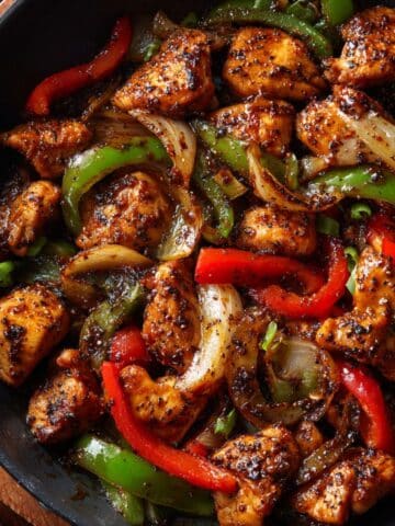 Juicy black pepper chicken stir-fry cooked in a skillet with glossy sauce, tender chicken bites, red and green bell peppers, and caramelized onions tossed in bold cracked black pepper seasoning.