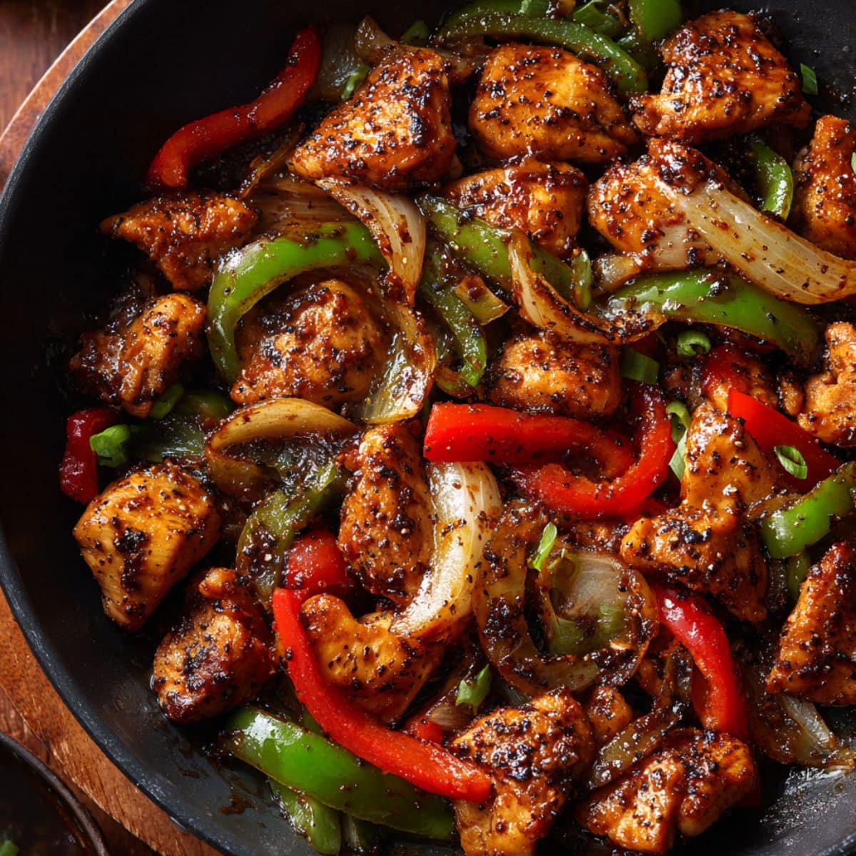 Juicy black pepper chicken stir-fry cooked in a skillet with glossy sauce, tender chicken bites, red and green bell peppers, and caramelized onions tossed in bold cracked black pepper seasoning.