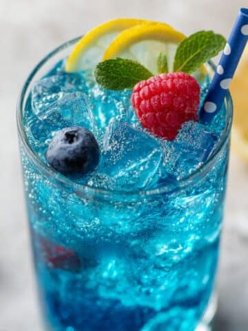glass of electric blue raspberry lemonade with fresh raspberries, blueberries, lemon slices, and mint, served over ice with a blue polka-dot straw on a white marble surface.