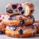 Close-up image of stacked baked blueberry donuts glazed with vanilla icing, one donut showing a soft bite revealing fluffy interior and fresh blueberries, styled with natural light for a glossy homemade look.