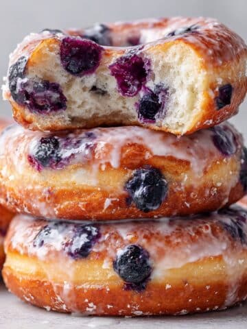 Close-up image of stacked baked blueberry donuts glazed with vanilla icing, one donut showing a soft bite revealing fluffy interior and fresh blueberries, styled with natural light for a glossy homemade look.