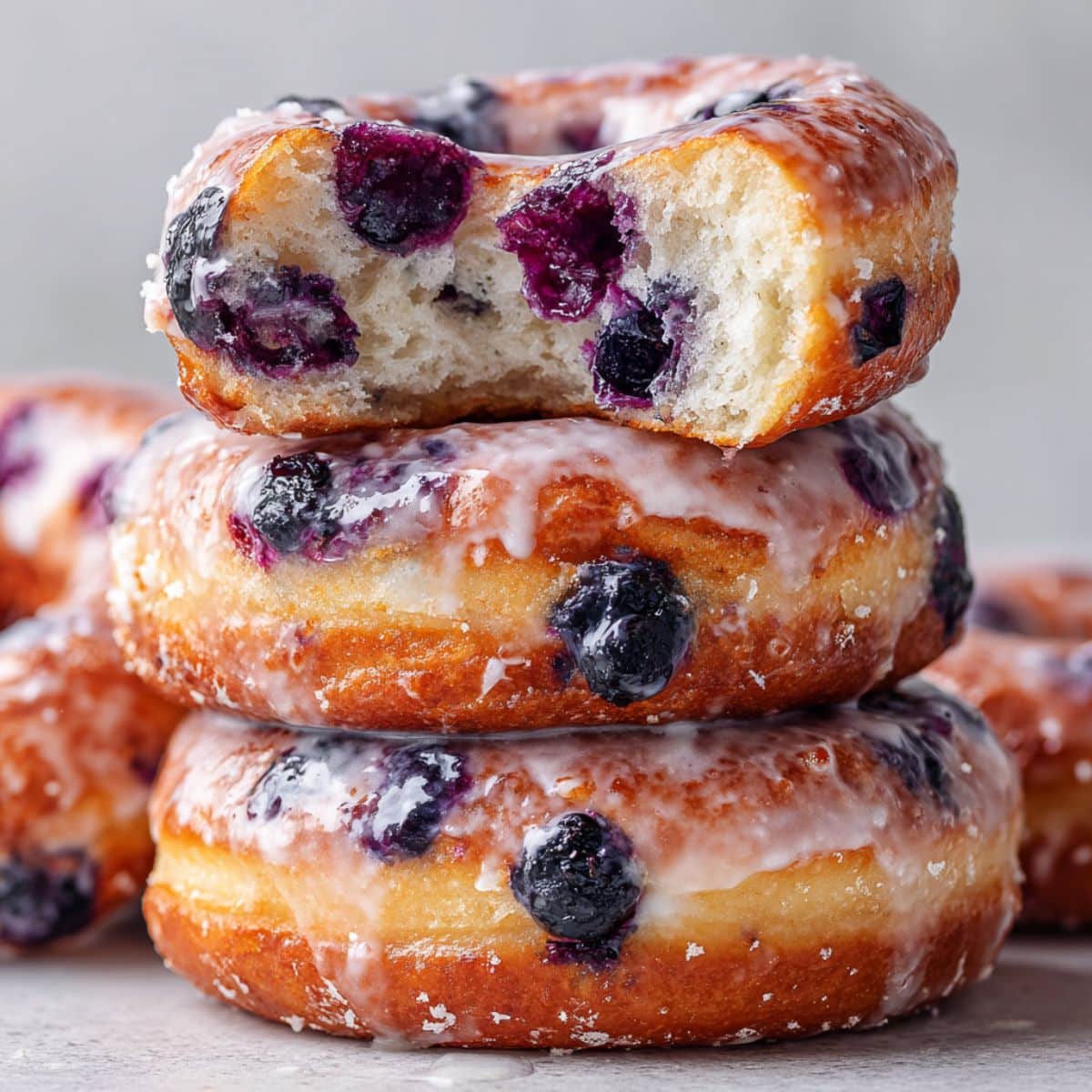 Close-up image of stacked baked blueberry donuts glazed with vanilla icing, one donut showing a soft bite revealing fluffy interior and fresh blueberries, styled with natural light for a glossy homemade look.