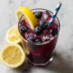 Refreshing homemade blueberry lemonade with ice, lemon wedges, and blueberries in clear glasses, served with striped straws on a rustic wooden surface.