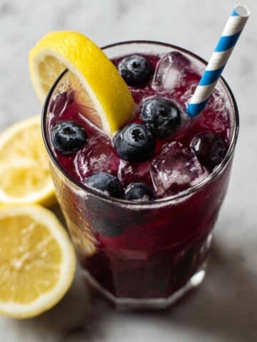Refreshing homemade blueberry lemonade with ice, lemon wedges, and blueberries in clear glasses, served with striped straws on a rustic wooden surface.
