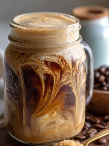 Side view of homemade brown sugar shaken espresso with layered swirl and frothy top in mason jar. Set on a linen towel with brown sugar and coffee beans.