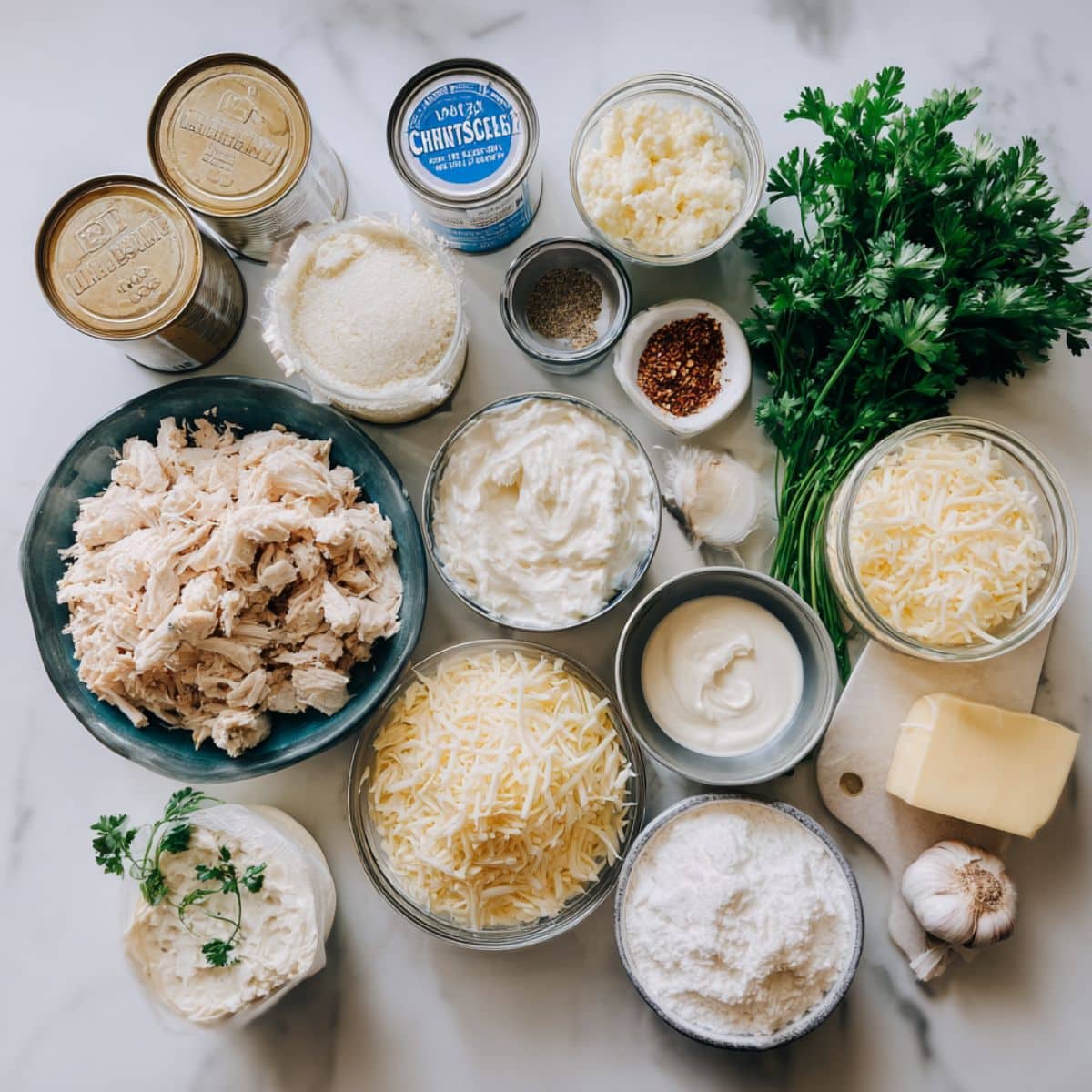 All ingredients for chicken alfredo monkey bread displayed on a white counter. Includes biscuit dough, chicken, cheeses, sauce, butter, and herbs.