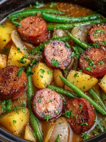 Hearty crockpot kielbasa and green beans with golden potatoes, caramelized onions, and smoked sausage slices, all coated in a savory broth and topped with fresh parsley for a rustic, homemade comfort dish.