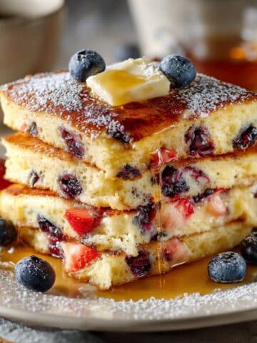 Close-up of a tall stack of square-cut sheet pan pancakes with golden fluffy layers, bursting with fresh blueberries and strawberries, topped with a pat of melting butter and drizzled maple syrup, served on a white plate with scattered berries.