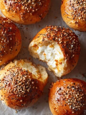 Golden brown everything bagel bites stuffed with creamy cheese, one torn open to reveal a rich, fluffy filling—close-up on parchment with seed mix in background