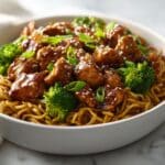 Glossy chicken teriyaki noodles served in a black bowl, topped with sesame seeds, green onions, and broccoli, with a rich brown sauce glistening over tender chicken pieces and stir-fried noodles.