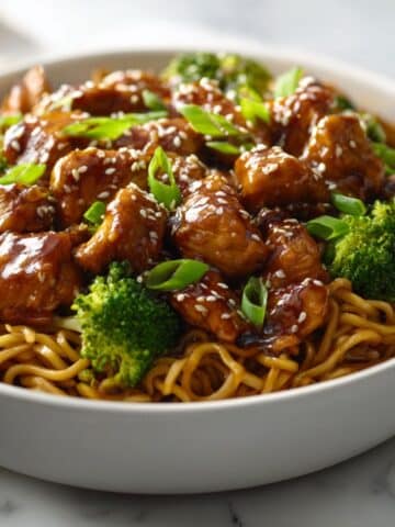 Glossy chicken teriyaki noodles served in a black bowl, topped with sesame seeds, green onions, and broccoli, with a rich brown sauce glistening over tender chicken pieces and stir-fried noodles.