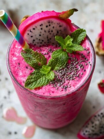 Vibrant pink dragon fruit smoothie with fresh mint garnish and dragon fruit slices in tall glass on white kitchen counter. Healthy pitaya smoothie with tropical flavors showing natural pink color from fresh dragon fruit with colorful straw and herbs.