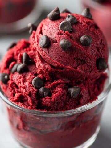 Edible red velvet cookie dough served in a clear glass cup, rich red color with a creamy soft texture and topped with semi-sweet chocolate chips. A homemade no-bake red velvet dessert recipe that’s safe to eat, indulgent, and perfect for chocolate lovers.