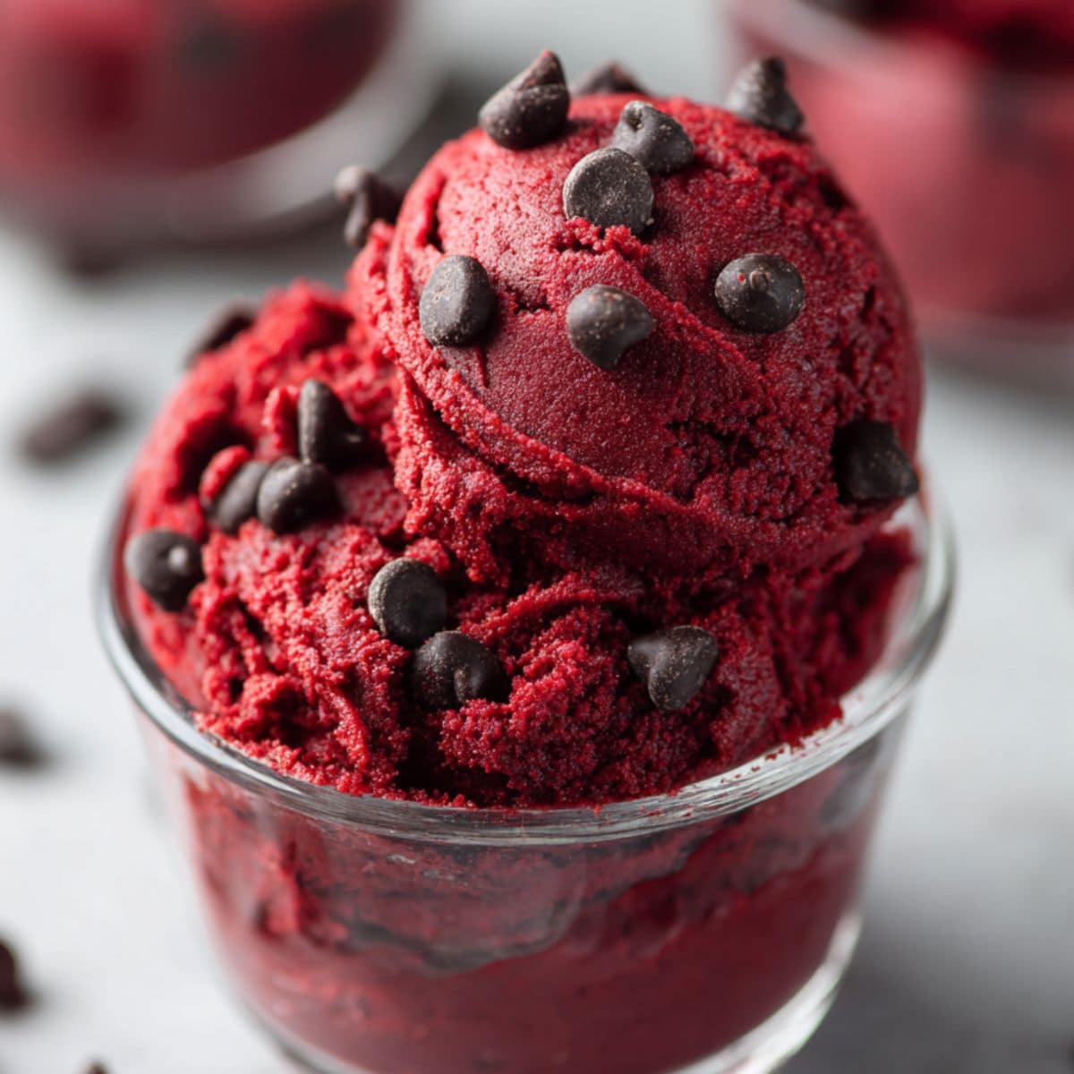 Edible red velvet cookie dough served in a clear glass cup, rich red color with a creamy soft texture and topped with semi-sweet chocolate chips. A homemade no-bake red velvet dessert recipe that’s safe to eat, indulgent, and perfect for chocolate lovers.