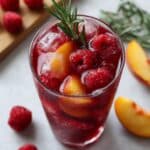 Deep red raspberry iced tea with fresh peach slices, raspberries and rosemary garnish in tall glasses on white kitchen counter.