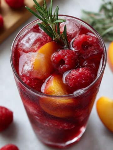 Deep red raspberry iced tea with fresh peach slices, raspberries and rosemary garnish in tall glasses on white kitchen counter.