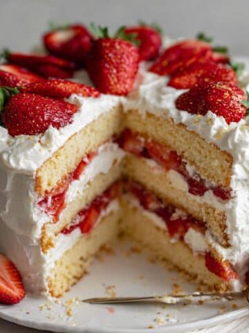 Layered strawberry shortcake cake with vanilla layers, fresh strawberries, and whipped cream frosting on white kitchen counter.