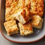 golden-baked butter swim biscuits in a white ceramic pan, cut into neat squares with lightly crisped tops and soft, fluffy centers, one piece pulled out showing a tender crumb, minimal melted butter pooling in the corner, styled on a neutral countertop with natural daylight.