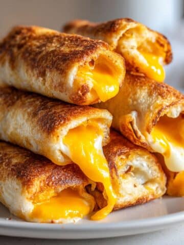 Golden brown grilled cheese roll-ups stacked on a white plate, made with crispy toasted bread and gooey melted American cheese oozing out of the ends.