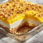 Side view of a golden lemon cream cheese dump cake in a glass baking dish, showing layers of buttery crumb crust, creamy filling, and glossy lemon topping with crumbles, baked to perfection for a rich, tangy, and homemade dessert.
