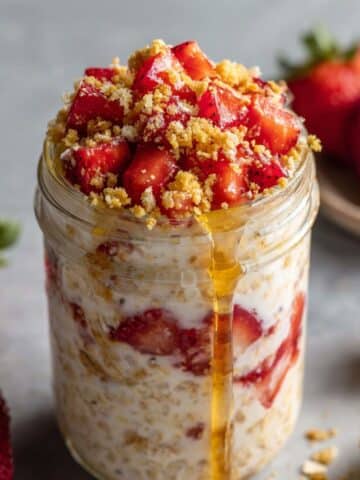Mason jar of strawberry shortcake overnight oats topped with fresh strawberries, graham crumbs, and honey drizzle on a light kitchen counter.