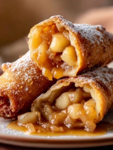 Golden brown apple pie egg rolls filled with warm cinnamon-spiced apple chunks, drizzled with glossy caramel sauce and dusted with powdered sugar for a crispy, gooey, and mouthwatering dessert.