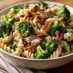 Creamy broccoli pasta salad with rotini noodles, fresh broccoli florets, crispy bacon, and red onion, served in a white bowl. The vibrant colors and creamy texture make it a refreshing and mouthwatering side dish