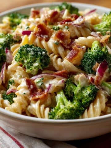 Creamy broccoli pasta salad with rotini noodles, fresh broccoli florets, crispy bacon, and red onion, served in a white bowl. The vibrant colors and creamy texture make it a refreshing and mouthwatering side dish
