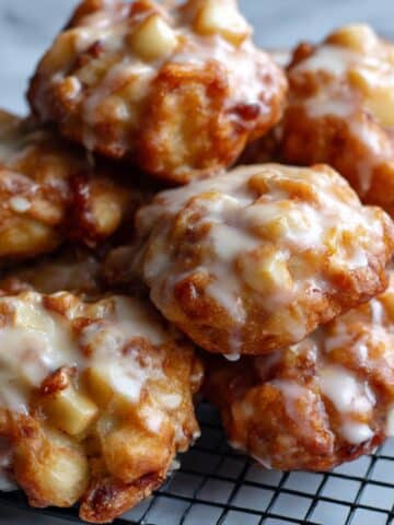 Close-up of golden brown homemade apple fritters clustered on a black cooling rack over a white marble countertop, coated in thick vanilla glaze with glossy drips and rustic texture.