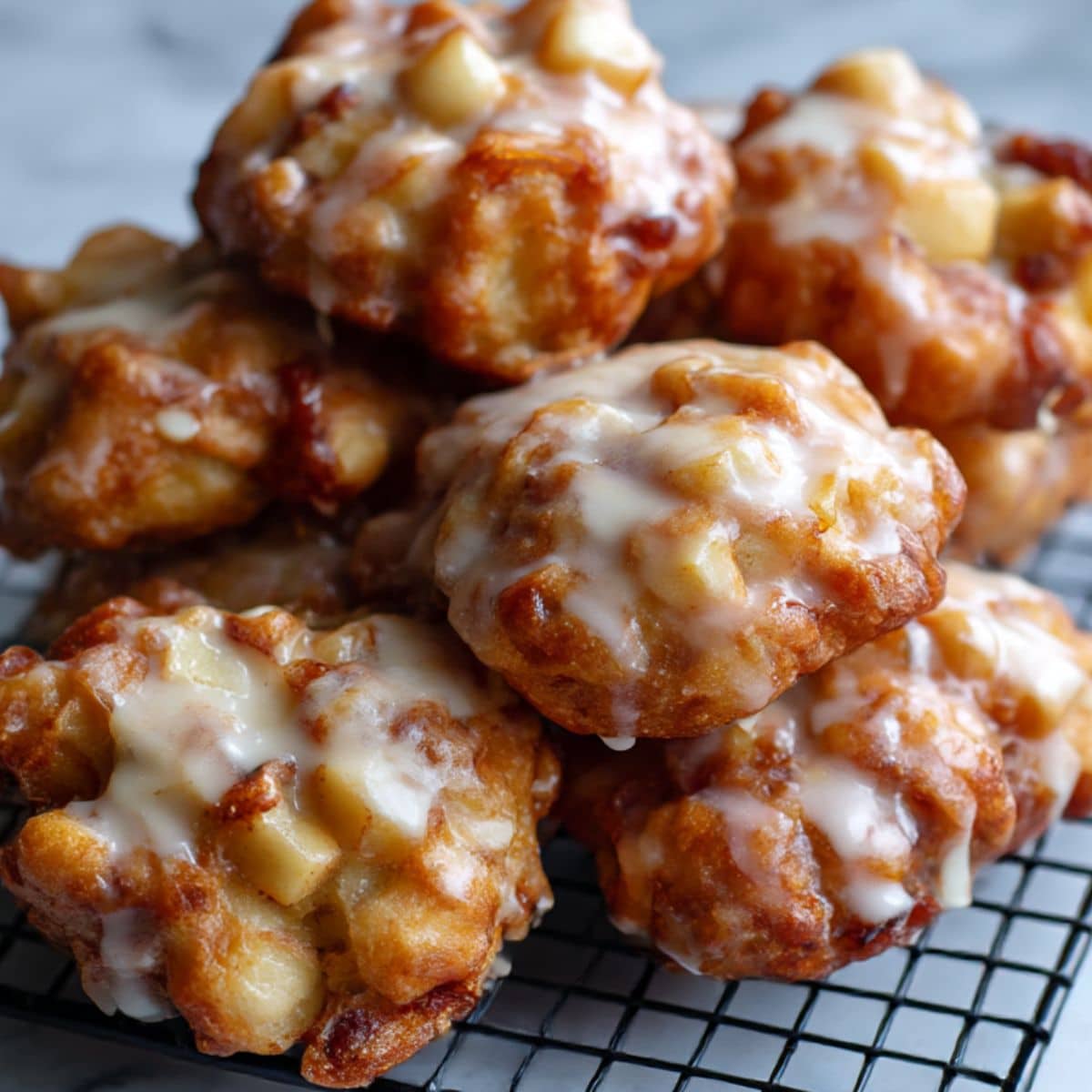 Close-up of golden brown homemade apple fritters clustered on a black cooling rack over a white marble countertop, coated in thick vanilla glaze with glossy drips and rustic texture.