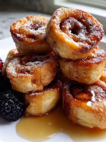 Golden homemade cinnamon roll french toast bites drizzled with syrup on a white kitchen counter. Crispy edges with soft cinnamon swirls inside.