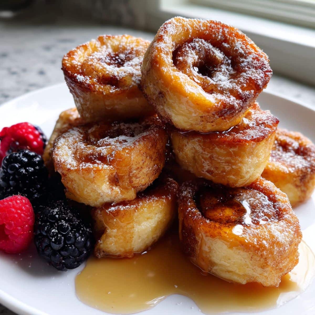 Golden homemade cinnamon roll french toast bites drizzled with syrup on a white kitchen counter. Crispy edges with soft cinnamon swirls inside.