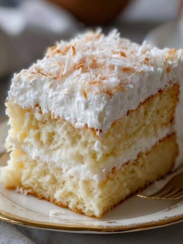 Slice of coconut cloud cake with fluffy whipped frosting and shredded coconut on a vintage plate. Real homemade texture with golden crumb and creamy layers.