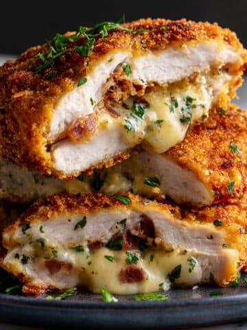 crispy golden Chicken Kiev cut in half, revealing a gooey, melted cheese and herb filling with bits of bacon. The breaded crust is perfectly browned and flecked with parsley, with cheese oozing onto a dark plate for an ultra-juicy, mouthwatering finish.