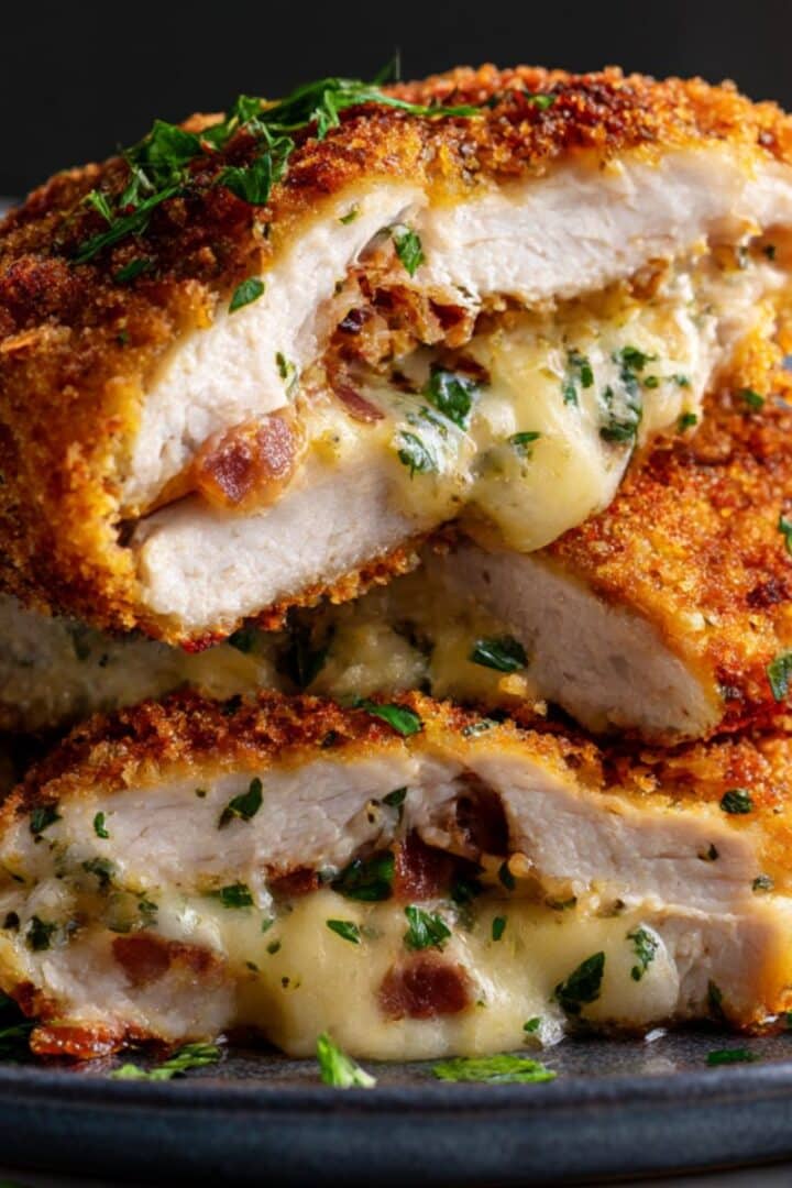 crispy golden Chicken Kiev cut in half, revealing a gooey, melted cheese and herb filling with bits of bacon. The breaded crust is perfectly browned and flecked with parsley, with cheese oozing onto a dark plate for an ultra-juicy, mouthwatering finish.