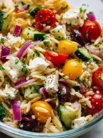 Overhead view of a vibrant Greek orzo salad filled with cherry tomatoes, cucumbers, black olives, red onions, feta cheese, and fresh parsley, all tossed in a light dressing and sprinkled with cracked black pepper.
